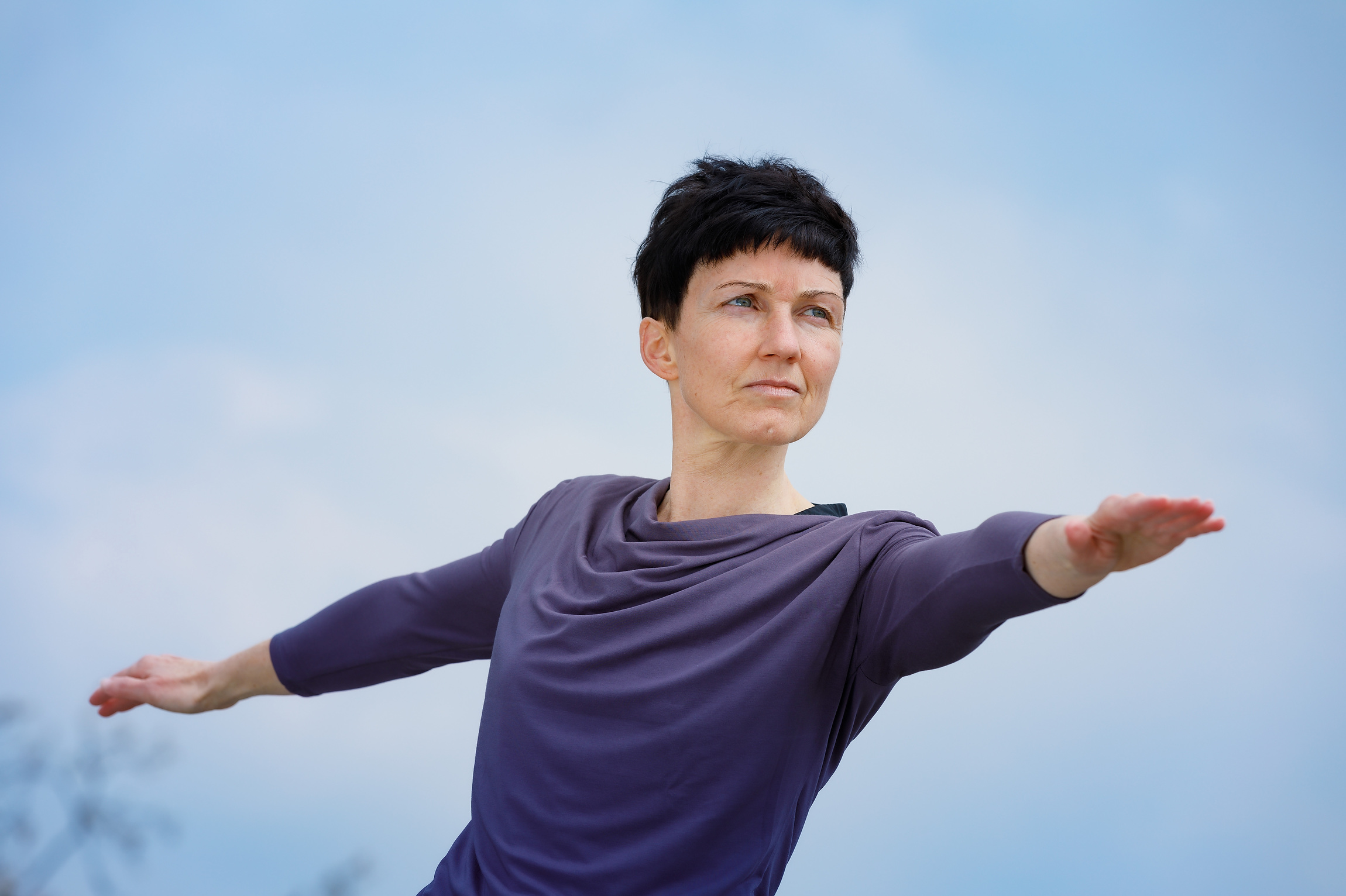 Porträt Sabine Docekal - schwarze kurze Haare, leuchtend blaue Augen, nimmt vor strahlend blauem Himmel die Kriegerpose ein.