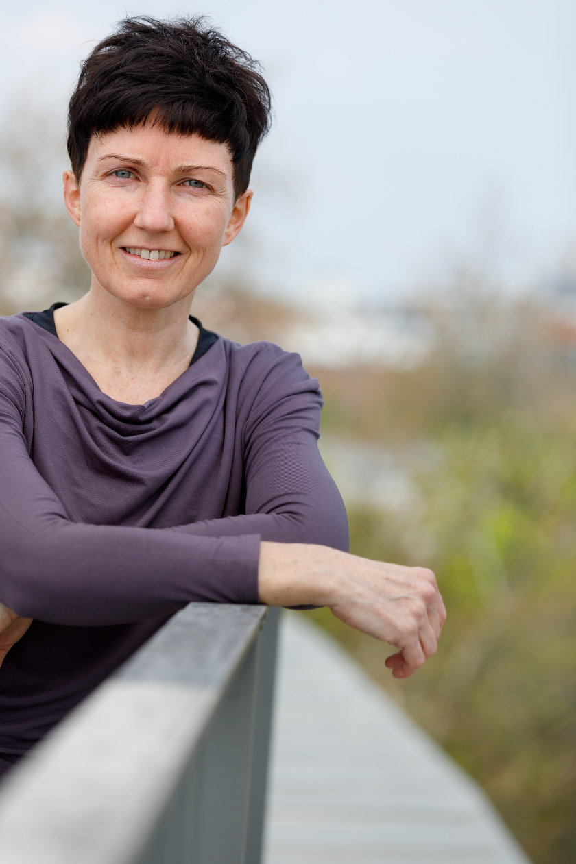 Porträt Sabine Docekal - schwarze kurze Haare, leuchtend blaue Augen, lächelt ernst während sie sich ans Geländer der Dachterrasse lehnt.