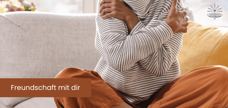 Eine Frau sitzt auf einem gemütlichen Sofa im Schneidersitz und umarmt sich selbst. Freundschaft mit dir.