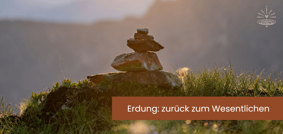 Nahaufnahme eines Kieseltürmchens im sonnenbeschienen Gras - dahinter sind Berge im Dunst zu erkennen.